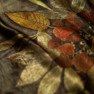 May include: A dark brown fabric scarf featuring a botanical print with detailed golden yellow and deep rust-colored leaf patterns. The textile has a soft, draped texture with visible veins on the leaves and a smooth, slightly reflective finish.