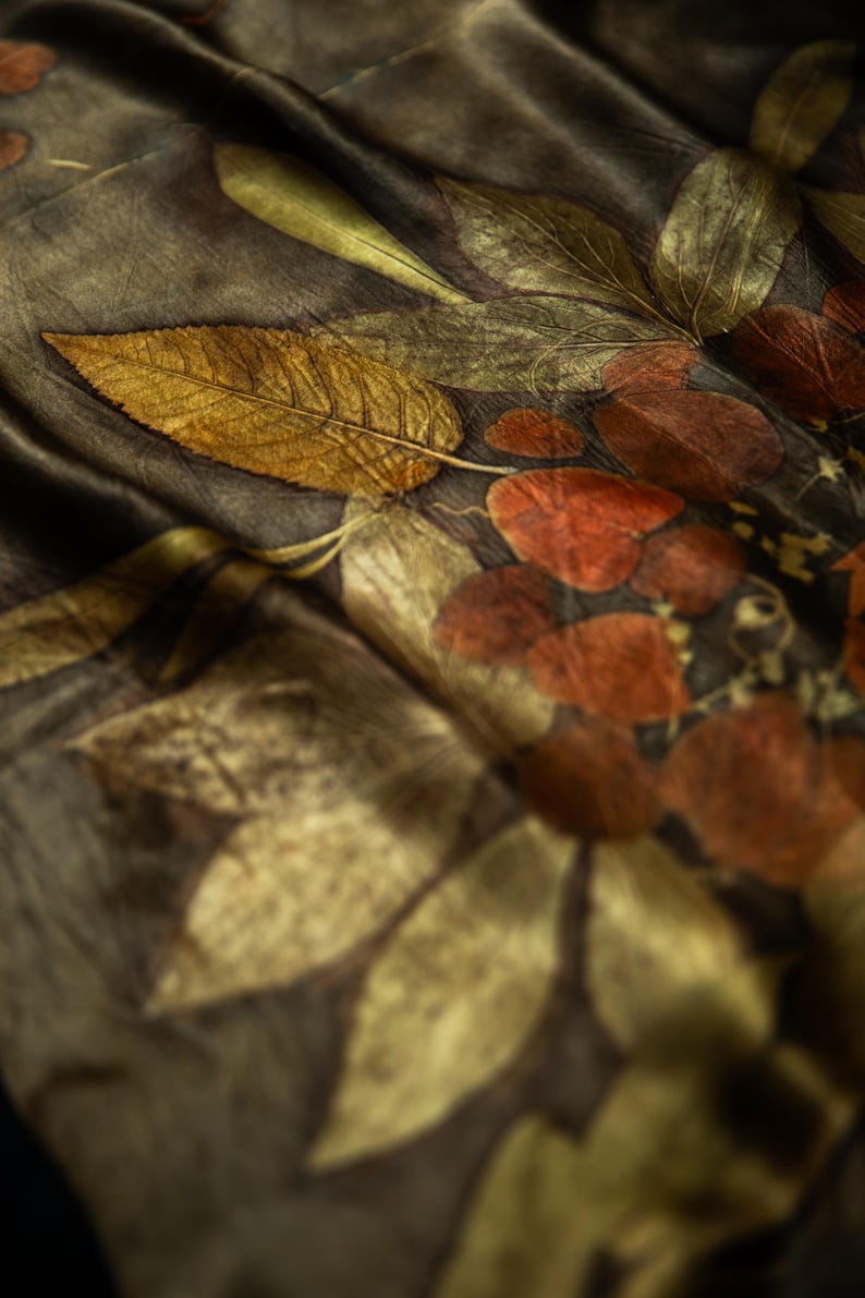 May include: A dark brown fabric scarf featuring a botanical print with detailed golden yellow and deep rust-colored leaf patterns. The textile has a soft, draped texture with visible veins on the leaves and a smooth, slightly reflective finish.