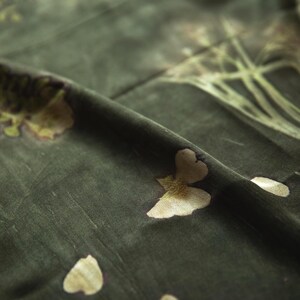 May include: A dark olive green fabric featuring a botanical print with light beige floral and leaf motifs. The material has a soft, matte texture with visible folds and creases that create depth across the surface of the textile.