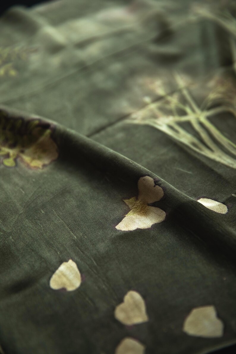 May include: A dark olive green fabric featuring a botanical print with light beige floral and leaf motifs. The material has a soft, matte texture with visible folds and creases that create depth across the surface of the textile.