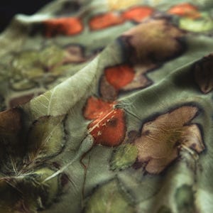 May include: A lightweight fabric scarf featuring a botanical print with a sage green background. The textile displays a repeating pattern of pressed leaves and foliage in shades of deep forest green, earthy brown, and vibrant burnt orange tones.