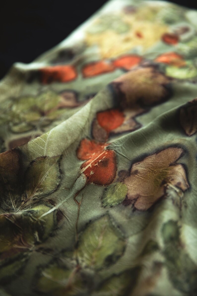 May include: A lightweight fabric scarf featuring a botanical print with a sage green background. The textile displays a repeating pattern of pressed leaves and foliage in shades of deep forest green, earthy brown, and vibrant burnt orange tones.