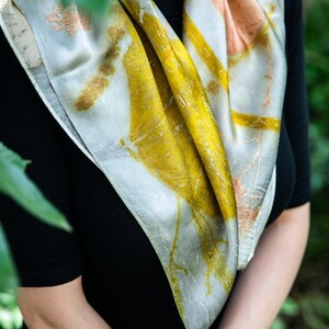 May include: A square silk scarf featuring a botanical print with vibrant yellow, soft peach, and muted grey tones. The fabric displays detailed leaf and plant imprints across the surface, creating a natural pattern that drapes around the neck.