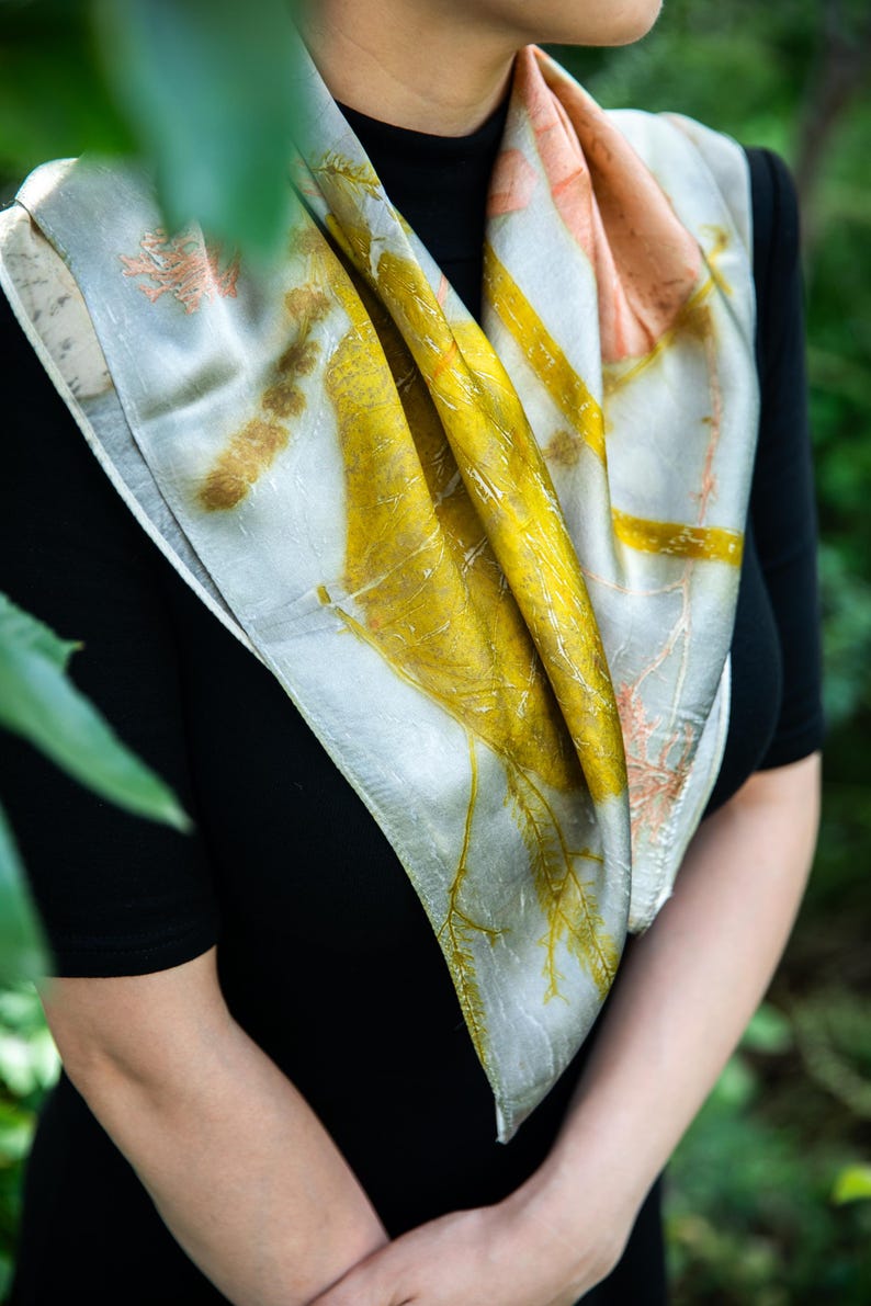 May include: A square silk scarf featuring a botanical print with vibrant yellow, soft peach, and muted grey tones. The fabric displays detailed leaf and plant imprints across the surface, creating a natural pattern that drapes around the neck.