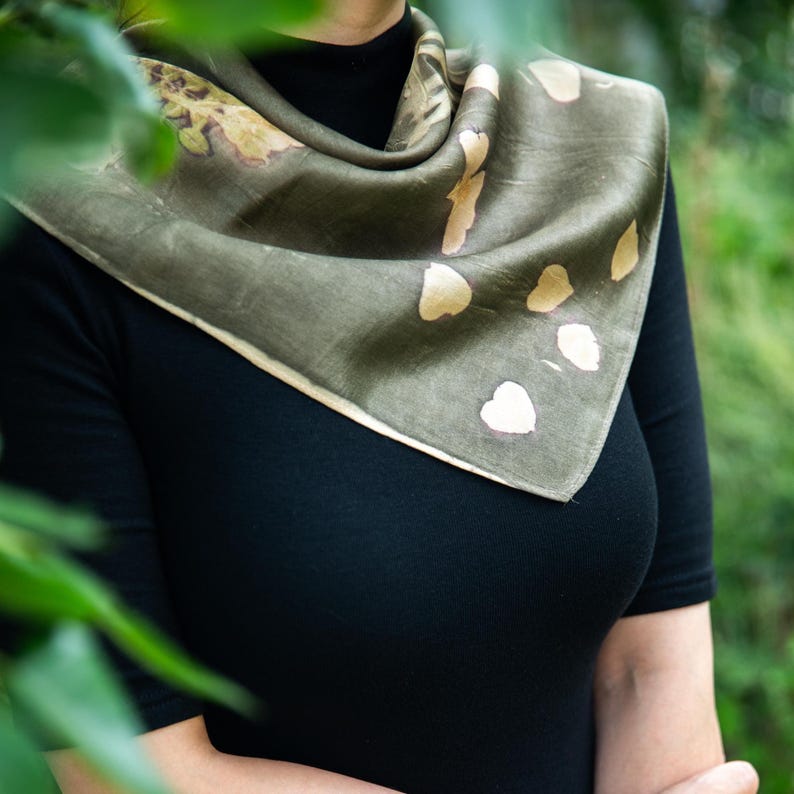 May include: A square scarf in a muted olive green shade featuring a botanical print with scattered light beige leaf and petal shapes. The scarf is draped around the neck, showcasing a smooth texture and a folded edge that reveals the fabric's drape.