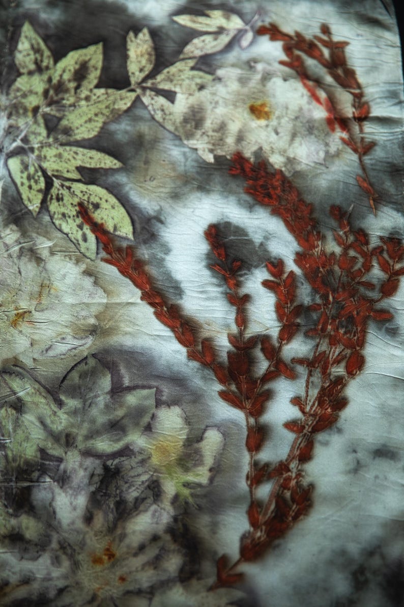 May include: A textile piece featuring a botanical print with dark gray and white marbled background tones. The fabric displays detailed imprints of light green leaves and vibrant rust-red sprigs, creating a natural, layered floral pattern across the surface.