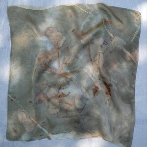 May include: A square scarf featuring a botanical print with muted sage green and gray tones. The fabric displays detailed imprints of leaves and thin branches in shades of rust orange and brown, creating a natural, layered pattern across the surface.