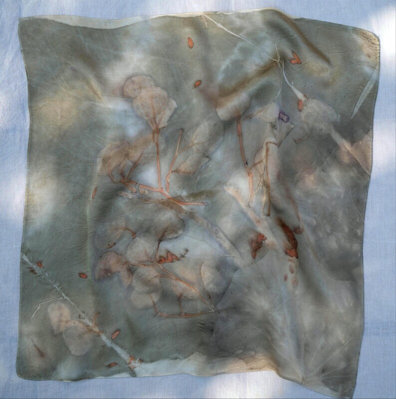 May include: A square scarf featuring a botanical print with muted sage green and gray tones. The fabric displays detailed imprints of leaves and thin branches in shades of rust orange and brown, creating a natural, layered pattern across the surface.