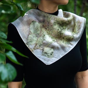 May include: A square scarf featuring a botanical print with green leaf and foliage patterns on a light, off-white background. The scarf is draped around the neck, displaying organic, earthy textures and natural plant imprints across the fabric.