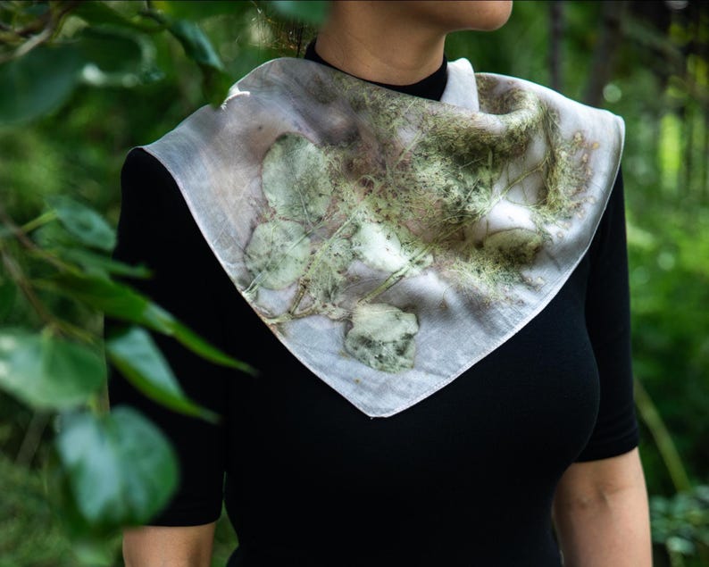 May include: A square scarf featuring a botanical print with green leaf and foliage patterns on a light, off-white background. The scarf is draped around the neck, displaying organic, earthy textures and natural plant imprints across the fabric.