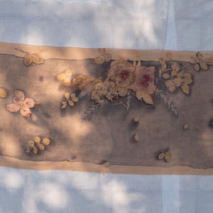 May include: A rectangular scarf featuring a botanical print with muted grey and brown tones. The fabric displays a central panel decorated with pressed flower and leaf silhouettes in shades of beige and soft pink, framed by a solid tan border.