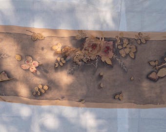 Puede incluir: Una bufanda rectangular con un estampado botánico en tonos grises y marrones apagados. La tela muestra un panel central decorado con siluetas de flores y hojas prensadas en tonos beige y rosa suave, enmarcado por un borde de color canela.
