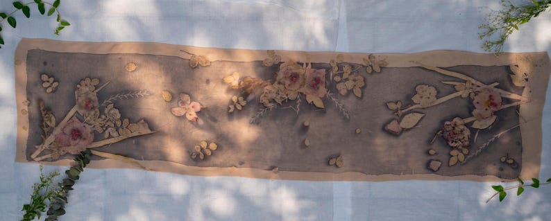 May include: A rectangular scarf featuring a botanical print with muted grey and brown tones. The fabric displays a central panel decorated with pressed flower and leaf silhouettes in shades of beige and soft pink, framed by a solid tan border.