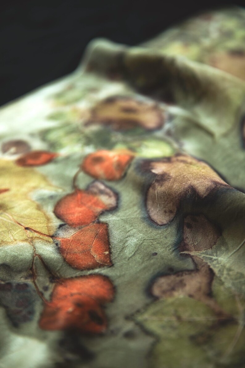 May include: A piece of fabric featuring a botanical print with a soft sage green background. The textile displays a pattern of pressed leaves in shades of burnt orange, deep brown, and muted yellow, creating a natural, organic appearance.