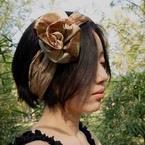 Pu&ograve; includere: Una fascia per capelli in raso color marrone dorato con un grande dettaglio a forma di rosa scolpita in tessuto sul lato. L'accessorio ha una finitura liscia e lucente, avvolto attorno alla testa, con una texture drappeggiata e un tono caldo.