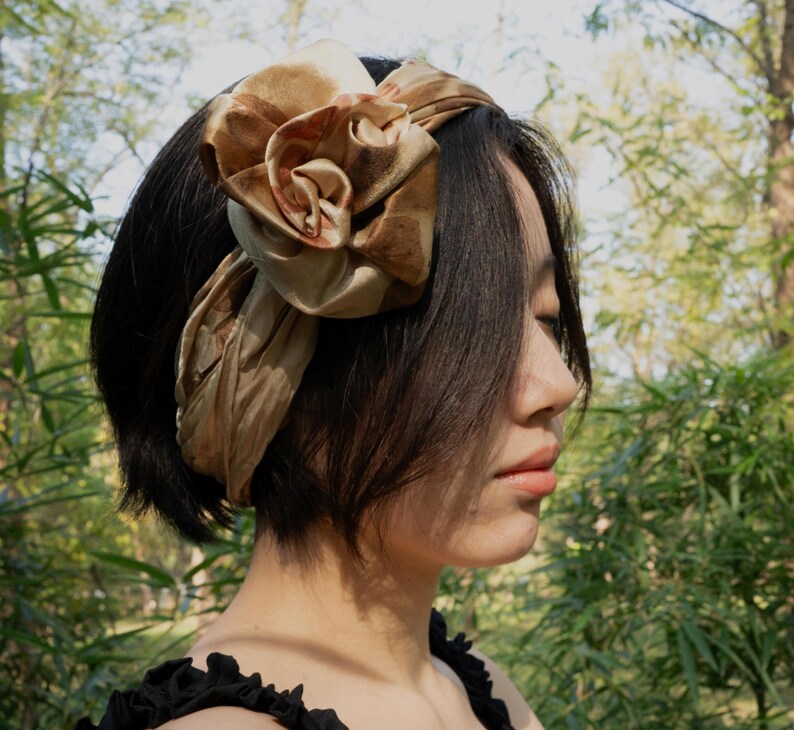 Pu&ograve; includere: Una fascia per capelli in raso color marrone dorato con un grande dettaglio a forma di rosa scolpita in tessuto sul lato. L'accessorio ha una finitura liscia e lucente, avvolto attorno alla testa, con una texture drappeggiata e un tono caldo.