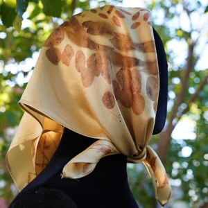 May include: A square silk scarf in a pale cream color featuring a botanical print with brown leaf and petal patterns. The scarf is tied in a knot at the side, displaying a smooth texture and a natural, earthy aesthetic with visible plant imprints.