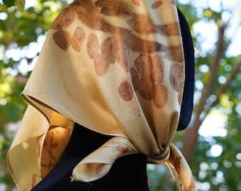 Puede incluir: Un pañuelo cuadrado de seda en color crema pálido con un estampado botánico de hojas y pétalos marrones. El pañuelo está anudado en el lateral, mostrando una textura suave y una estética natural con impresiones de plantas claramente visibles.