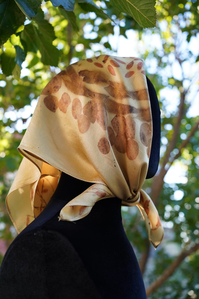 Pu&ograve; includere: Un foulard quadrato in seta color crema pallido con una stampa botanica di foglie e petali marroni. Il foulard &egrave; annodato lateralmente e presenta una consistenza liscia e un'estetica naturale con impronte di piante chiaramente visibili.