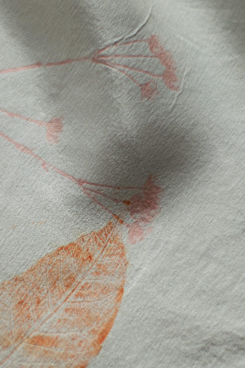 May include: A light gray fabric featuring a botanical print with delicate pink flower stems and a prominent orange leaf. The material has a visible, slightly textured weave, and the printed elements appear as soft, organic impressions.