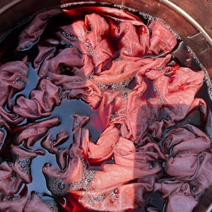 May include: A metal pot filled with a dark red liquid dye and submerged fabric. The fabric is bunched and folded, showing varying shades of pink and mauve as it absorbs the deep crimson pigment. Small bubbles are visible on the surface of the liquid.