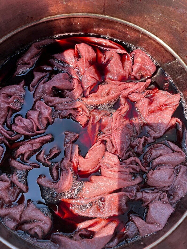 May include: A metal pot filled with a dark red liquid dye and submerged fabric. The fabric is bunched and folded, showing varying shades of pink and mauve as it absorbs the deep crimson pigment. Small bubbles are visible on the surface of the liquid.