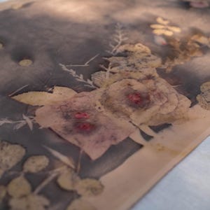 May include: A textile with a floral design. The fabric features a muted color palette with a dark gray background and light beige edges. The floral pattern includes roses and other botanical elements in shades of beige, pink, and yellow.