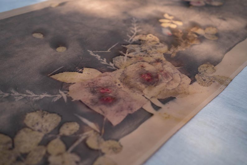 May include: A textile with a floral design. The fabric features a muted color palette with a dark gray background and light beige edges. The floral pattern includes roses and other botanical elements in shades of beige, pink, and yellow.
