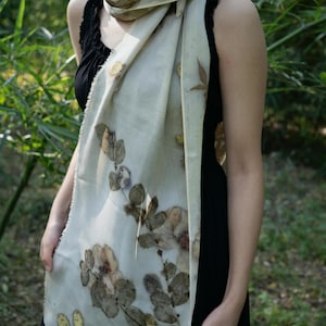 May include: A rectangular off-white scarf featuring a botanical print of dark brown and muted green leaves and flowers. The scarf has a lightweight, semi-sheer texture with frayed edges and is draped around the neck over a black sleeveless top.