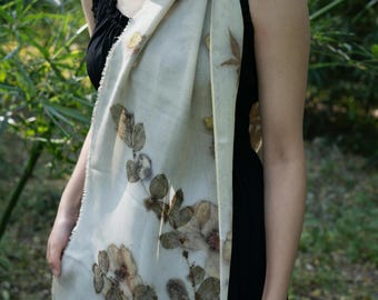 Puede incluir: Una bufanda rectangular de color blanco roto con un estampado botánico de hojas y flores en tonos marrón oscuro y verde apagado. La bufanda tiene una textura ligera y semitransparente con bordes deshilachados sobre una prenda negra.