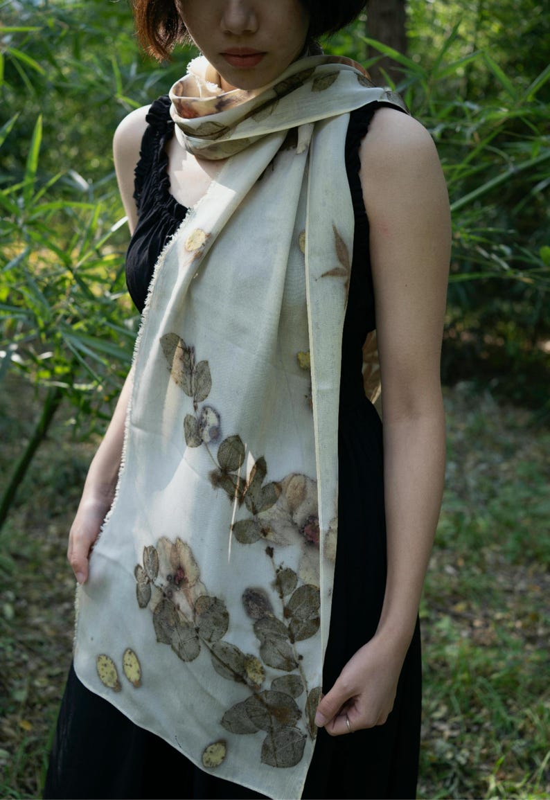 May include: A rectangular off-white scarf featuring a botanical print of dark brown and muted green leaves and flowers. The scarf has a lightweight, semi-sheer texture with frayed edges and is draped around the neck over a black sleeveless top.