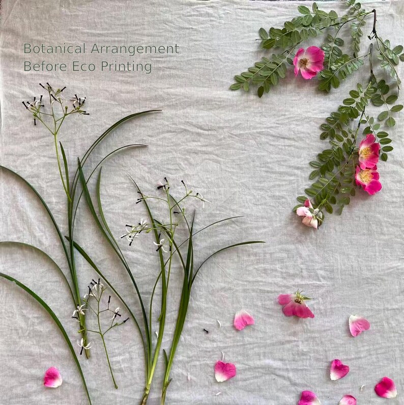 May include: A botanical arrangement on light gray fabric featuring green stems with small white flowers, a leafy branch with pink rose blossoms, and scattered pink petals. The text Botanical Arrangement Before Eco Printing is visible.