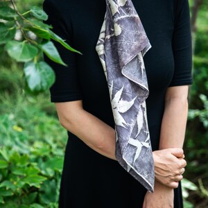 May include: A black short-sleeved dress paired with a rectangular scarf featuring a dark gray and white botanical leaf print. The scarf is tied around the neck and hangs down the front, displaying a repeating pattern of various foliage silhouettes.