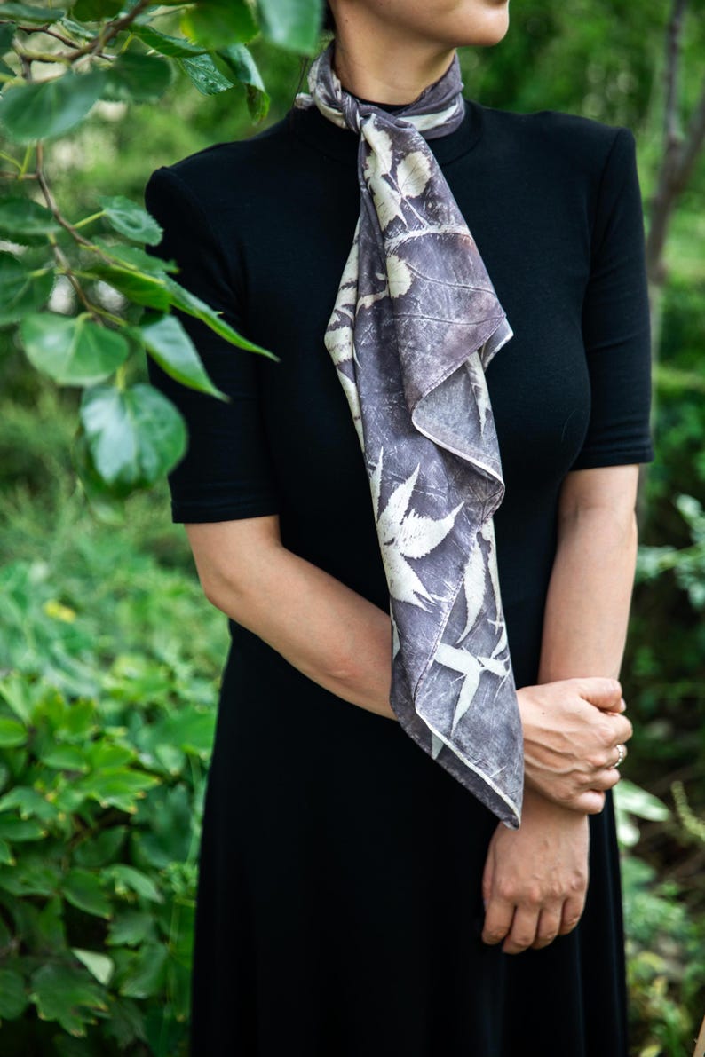 May include: A black short-sleeved dress paired with a rectangular scarf featuring a dark gray and white botanical leaf print. The scarf is tied around the neck and hangs down the front, displaying a repeating pattern of various foliage silhouettes.