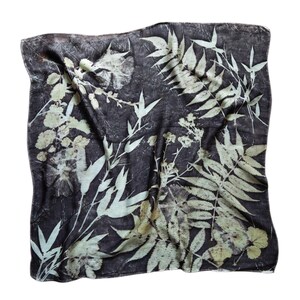 May include: A square scarf featuring a dark charcoal gray background with a botanical print of light cream-colored leaves, ferns, and floral sprigs. The fabric displays a natural, textured appearance with intricate plant silhouettes across the surface.
