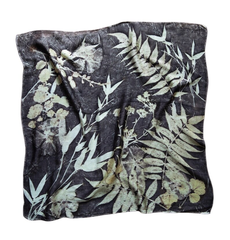 May include: A square scarf featuring a dark charcoal gray background with a botanical print of light cream-colored leaves, ferns, and floral sprigs. The fabric displays a natural, textured appearance with intricate plant silhouettes across the surface.