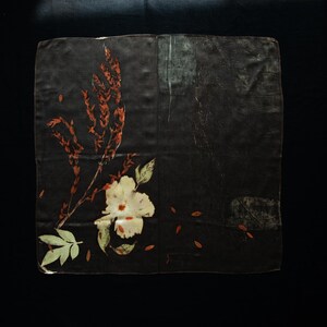 May include: A square dark brown fabric piece featuring a botanical print. The design includes a large cream-colored flower with green leaves at the bottom left, accompanied by tall, slender reddish-brown stalks and scattered small leaf-like shapes.