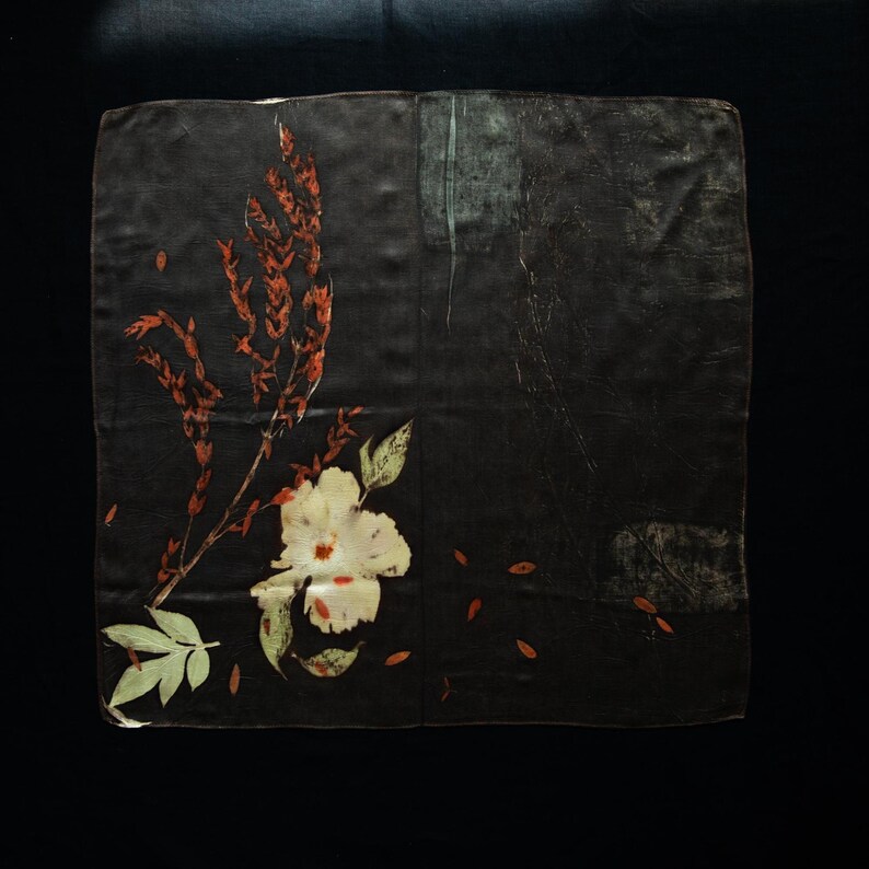 May include: A square dark brown fabric piece featuring a botanical print. The design includes a large cream-colored flower with green leaves at the bottom left, accompanied by tall, slender reddish-brown stalks and scattered small leaf-like shapes.