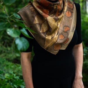 May include: A square silk scarf featuring an eco-printed botanical pattern with shades of brown, gold, and deep orange. The design displays various leaf shapes and circular motifs arranged in a border, draped over a solid black short-sleeved top.