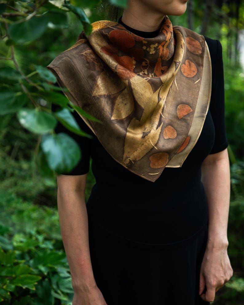 May include: A square silk scarf featuring an eco-printed botanical pattern with shades of brown, gold, and deep orange. The design displays various leaf shapes and circular motifs arranged in a border, draped over a solid black short-sleeved top.