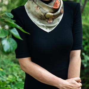 May include: A square silk scarf featuring a botanical print with muted green, brown, and orange tones. The scarf is folded into a triangle and tied around the neck, displaying organic leaf and floral patterns across the fabric surface.