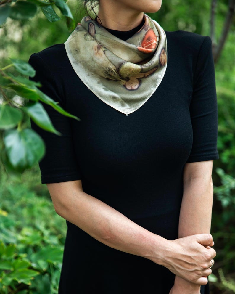 May include: A square silk scarf featuring a botanical print with muted green, brown, and orange tones. The scarf is folded into a triangle and tied around the neck, displaying organic leaf and floral patterns across the fabric surface.
