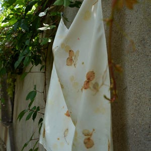 May include: A rectangular off-white fabric cloth featuring a botanical print with scattered brown and tan leaf and berry patterns. The textile is draped vertically, displaying a smooth surface texture and a consistent, repeating natural plant motif.