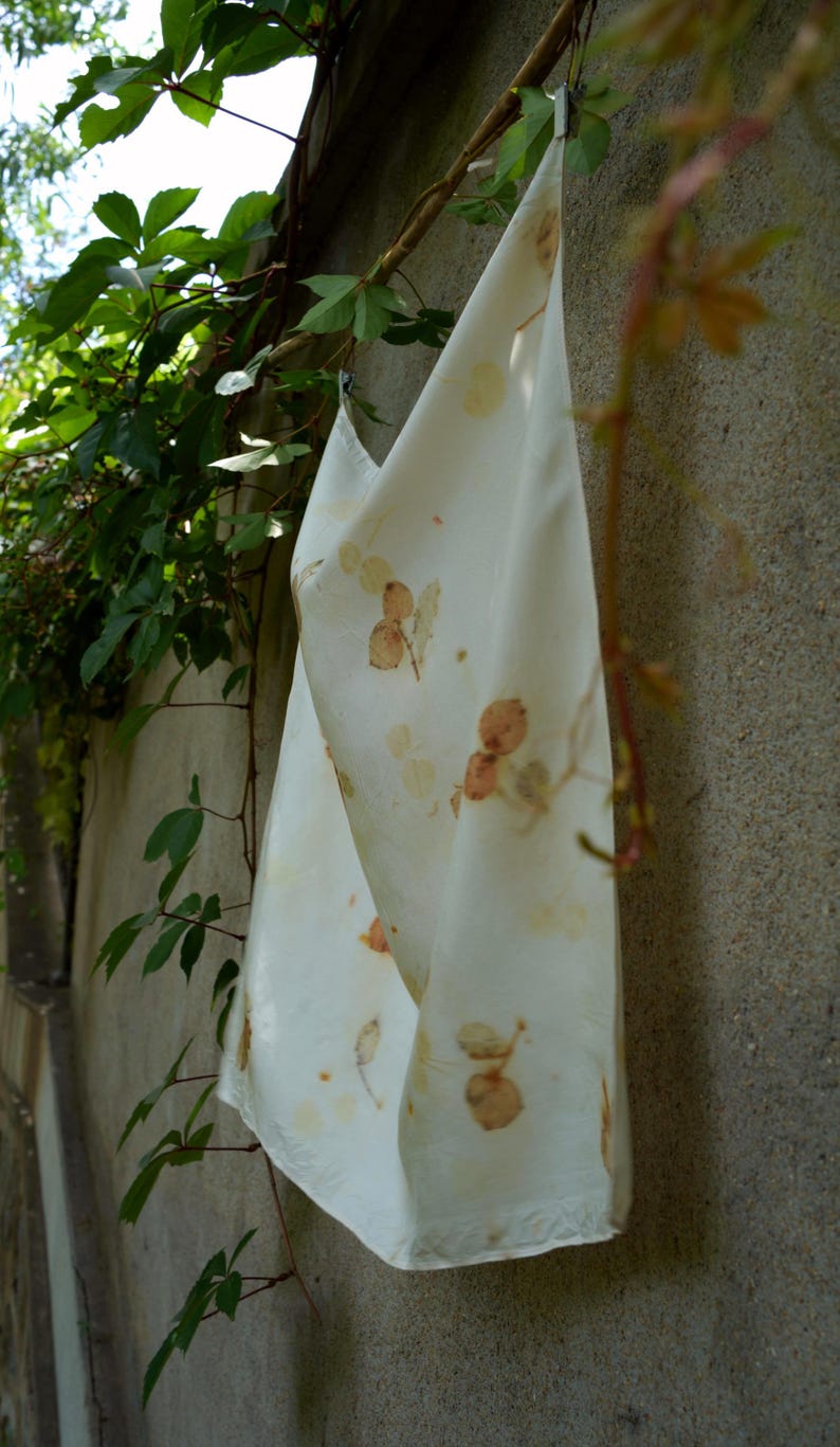 May include: A rectangular off-white fabric cloth featuring a botanical print with scattered brown and tan leaf and berry patterns. The textile is draped vertically, displaying a smooth surface texture and a consistent, repeating natural plant motif.