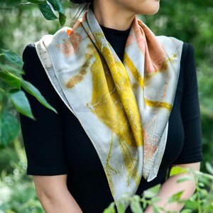 May include: A square silk scarf featuring botanical prints in shades of mustard yellow, soft peach, and muted grey. The fabric displays intricate, organic leaf and stem patterns across its surface, creating a natural, multi-colored design.
