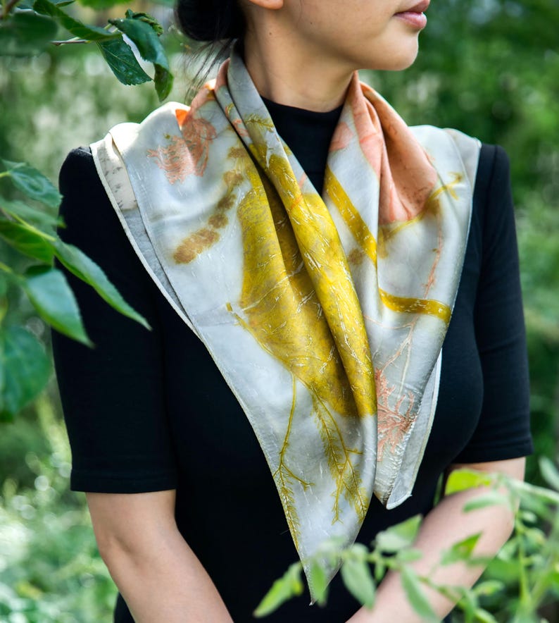 May include: A square silk scarf featuring botanical prints in shades of mustard yellow, soft peach, and muted grey. The fabric displays intricate, organic leaf and stem patterns across its surface, creating a natural, multi-colored design.