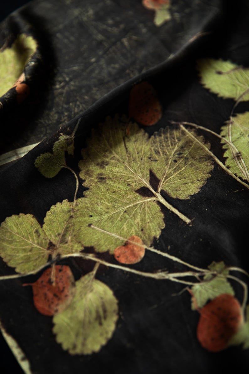 May include: A black fabric featuring a botanical print of green leaves and small reddish-brown circular shapes. The leaves display detailed veins and serrated edges, scattered across the dark surface in a natural, organic arrangement.