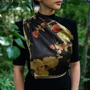 May include: A dark brown silk scarf featuring a botanical print of golden-yellow strawberry leaves and reddish-orange berries. The scarf is draped over a black short-sleeved top, showcasing a smooth texture and a repeating natural plant pattern.