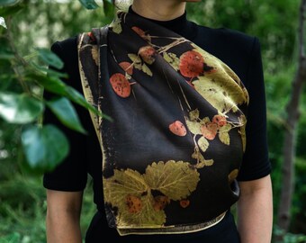 Puede incluir: Un pañuelo de seda marrón oscuro con un estampado botánico de hojas de fresa amarillo dorado y bayas rojo anaranjado. El pañuelo está drapeado sobre una camiseta negra de manga corta, mostrando una textura suave y un patrón vegetal natural.