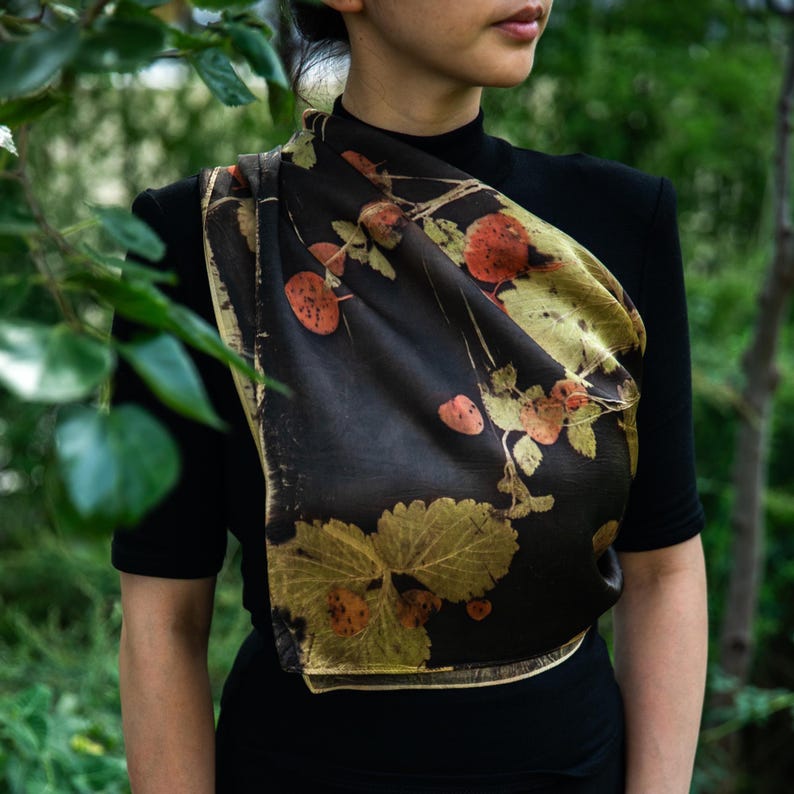 May include: A dark brown silk scarf featuring a botanical print of golden-yellow strawberry leaves and reddish-orange berries. The scarf is draped over a black short-sleeved top, showcasing a smooth texture and a repeating natural plant pattern.